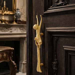 May include: A gold-colored door handle with a unique design, featuring a stylized figure reaching upwards. The handle is mounted on a dark brown wooden door, with intricate paneling and decorative elements. The overall aesthetic is elegant and artistic.