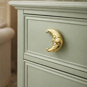 May include: A sage green dresser drawer with a gold-coloured crescent moon-shaped handle. The handle has a smiling face on the moon. The dresser has a classic design.