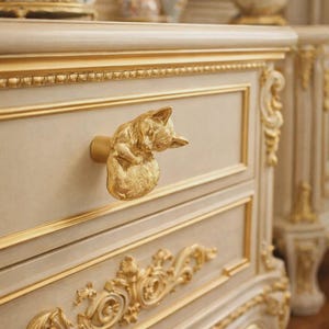 Może przedstawiać: Kremowa komoda ze złotymi wykończeniami i złotą gałką w kształcie kota. Komoda ma bogate rzeźbienia i klasyczny design. Obraz przedstawia zbliżenie szuflady i gałki w kształcie kota.