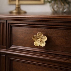 Può includere: Primo piano di un cassetto di una cassettiera in legno marrone scuro con una manopola dorata a forma di fiore. La manopola ha un design dettagliato e strutturato. La cassettiera fa parte di un set di mobili per la camera da letto.