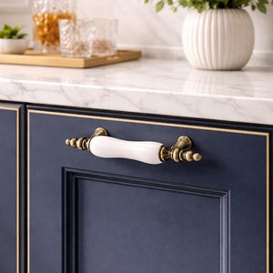 May include: Close-up of a navy blue cabinet door with a white and brass handle. The handle has a white ceramic center and brass accents. The cabinet door has a gold trim. A white marble countertop is above the cabinet.