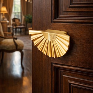 Peut inclure: Une poignée de porte dorée avec un design en forme d'éventail. La poignée est montée sur une porte en bois brun foncé avec des panneaux. Le design de la poignée présente une série de lignes rayonnantes et un dessus semi-circulaire.