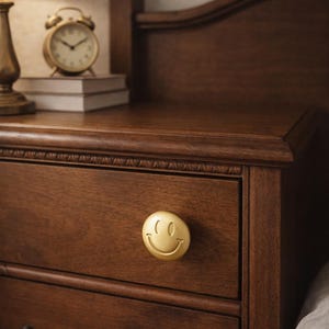 Puede incluir: Primer plano de una mesita de noche de madera oscura con un pomo dorado con forma de cara sonriente. Un reloj despertador dorado y una lámpara son visibles en el fondo. La madera es de color marrón oscuro.