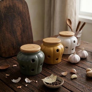 Può includere: Tre contenitori in ceramica per l'aglio, verdi, gialli e bianchi, con coperchi in legno, sono su un tavolo di legno. Ogni contenitore ha diversi fori di ventilazione circolari. Sono visibili anche spicchi d'aglio, una piccola ciotola di spezie e un tagliere di legno.