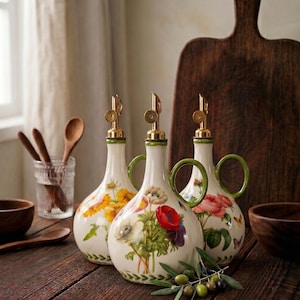 May include: Three decorative ceramic oil bottles with floral designs. Each bottle has a gold spout, green handle, and a different flower pattern: sunflowers, anemones, and roses. The bottles are on a wooden table with olive branches.