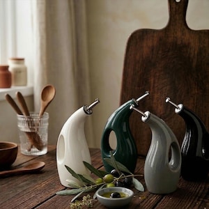 May include: Four ceramic oil dispensers in white, green, grey, and black, with silver spouts, sit on a wooden table. A small bowl of olives and olive oil, wooden spoons, and a cutting board are also visible, suggesting a kitchen or dining setting.