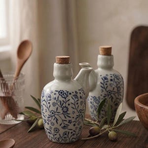 Puede incluir: Dos botellas de aceite de cerámica blanca con diseños florales azules y tapones de corcho. Las botellas tienen un pico vertedor y se exhiben sobre una superficie de madera con ramas de olivo y aceitunas.
