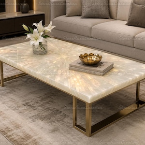 May include: A rectangular coffee table featuring a white, translucent, crystalline stone top with a sunburst pattern and a polished gold-tone metal frame. A clear glass vase with white lilies, a gold bowl, and two stacked books sit on the surface.