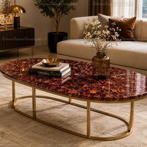 Puede incluir: Una mesa de centro ovalada con un tablero de piedra ágata de color rojo intenso pulido con un patrón de mosaico de piedras translúcidas. La mesa está sostenida por una estructura de metal fino en tono dorado con una base elíptica abierta.