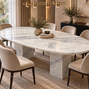Puede incluir: Una mesa de comedor ovalada con un tablero de mármol blanco con vetas grises, sostenida por dos gruesos pedestales rectangulares de mármol. La mesa está rodeada por seis sillas tapizadas con asientos de tela beige y patas de madera marrón oscuro.