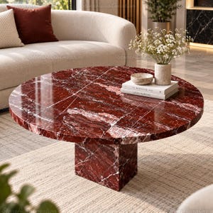 May include: A round coffee table featuring a deep red marble surface with white veining and a matching square pedestal base. On the table sits a small stone bowl, a white vase with white flowers, and two stacked books labeled The Kinfolk and Louis Vuitton.