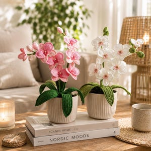 May include: Two crochet orchid plants in cream-colored pots. One orchid has pink flowers, the other has white flowers. Both have green leaves and are sitting on a wooden table. The table also has books and a candle.