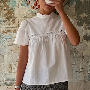 May include: A white short-sleeved blouse featuring a ruffled high neckline and a horizontal band of intricate eyelet lace detailing across the chest, paired with black and white checkered shorts that have a subtle ruffled hemline along the bottom.
