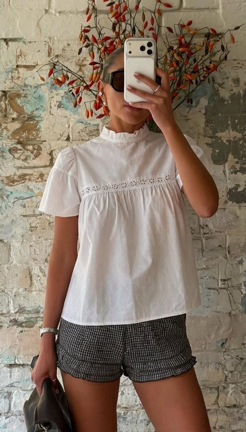 May include: A white short-sleeved blouse featuring a ruffled high neckline and a horizontal band of intricate eyelet lace detailing across the chest, paired with black and white checkered shorts that have a subtle ruffled hemline along the bottom.
