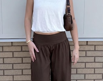 May include: A sleeveless white top paired with high-waisted, wide-leg brown pants. A small brown purse hangs from the shoulder. The outfit is accessorized with a gold bracelet and a delicate necklace. The background is a brick wall.