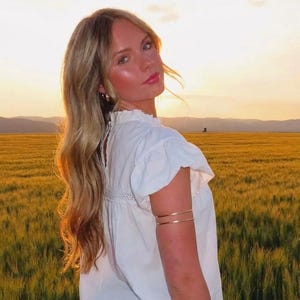Puede incluir: Una persona con una blusa blanca con mangas con volantes y cuello alto, combinada con pantalones cortos a cuadros. Lleva brazaletes dorados y una pulsera. El fondo presenta un campo dorado y una puesta de sol.