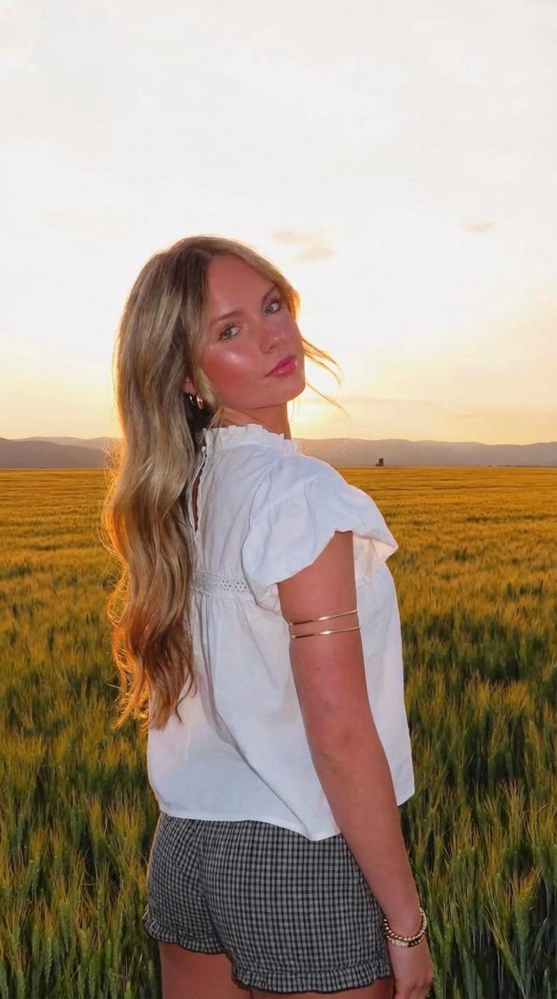 Puede incluir: Una persona con una blusa blanca con mangas con volantes y cuello alto, combinada con pantalones cortos a cuadros. Lleva brazaletes dorados y una pulsera. El fondo presenta un campo dorado y una puesta de sol.