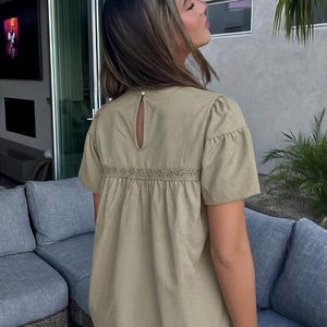 Puede incluir: Una blusa verde oliva claro con mangas cortas y un detalle de ojo de cerradura en la espalda. La blusa presenta una banda bordada decorativa en la secci&oacute;n media y se combina con vaqueros azules. La blusa tiene un corte holgado.