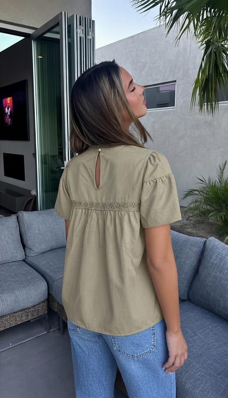 Puede incluir: Una blusa verde oliva claro con mangas cortas y un detalle de ojo de cerradura en la espalda. La blusa presenta una banda bordada decorativa en la secci&oacute;n media y se combina con vaqueros azules. La blusa tiene un corte holgado.