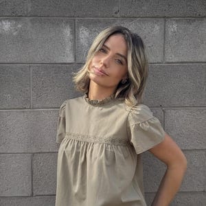 Puede incluir: Una blusa verde oliva claro con mangas cortas y cuello con volantes. La blusa tiene un detalle de encaje en el pecho y se combina con vaqueros azul claro y un cintur&oacute;n marr&oacute;n. El fondo es una pared de ladrillos gris.