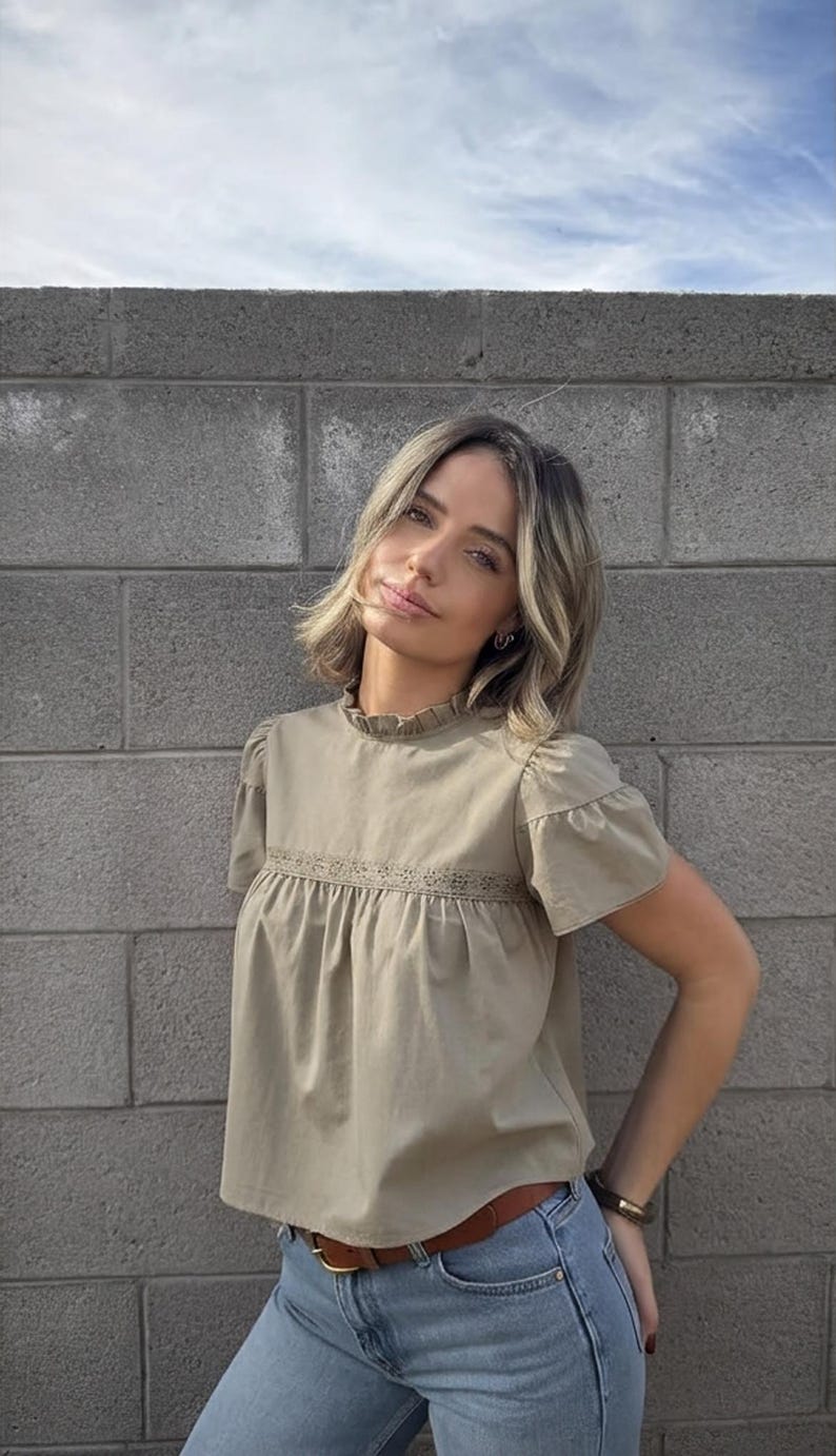 Puede incluir: Una blusa verde oliva claro con mangas cortas y cuello con volantes. La blusa tiene un detalle de encaje en el pecho y se combina con vaqueros azul claro y un cintur&oacute;n marr&oacute;n. El fondo es una pared de ladrillos gris.