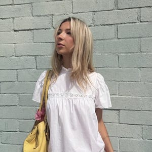 Puede incluir: Una mujer con una blusa blanca con detalles de encaje y una minifalda rosa a rayas. Lleva un bolso amarillo con una flor rosa. El fondo es una pared de ladrillos azul claro.