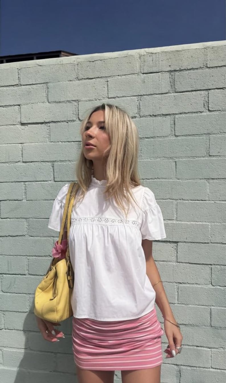 Puede incluir: Una mujer con una blusa blanca con detalles de encaje y una minifalda rosa a rayas. Lleva un bolso amarillo con una flor rosa. El fondo es una pared de ladrillos azul claro.