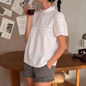 May include: A white short-sleeved blouse with a ruffled high neckline and a horizontal lace insert across the chest, paired with black and white checkered shorts featuring a ruffled hemline and side pockets, worn with large black oval earrings.