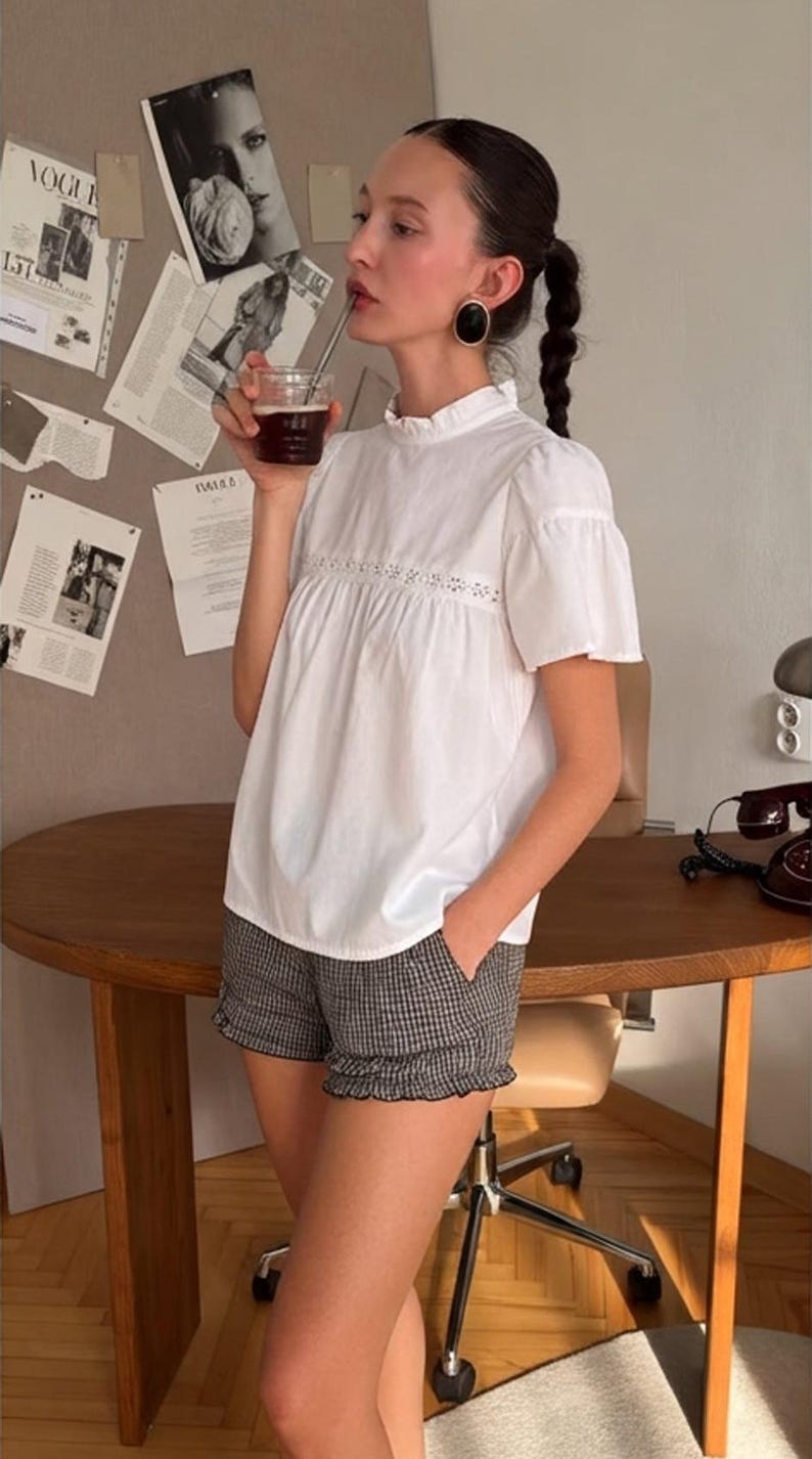 May include: A white short-sleeved blouse with a ruffled high neckline and a horizontal lace insert across the chest, paired with black and white checkered shorts featuring a ruffled hemline and side pockets, worn with large black oval earrings.