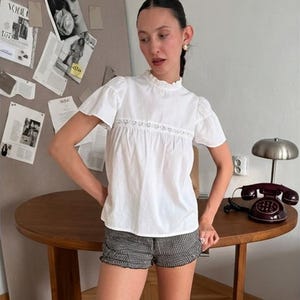 May include: A white short-sleeved blouse featuring a high ruffled collar and a horizontal lace insert across the chest, paired with black and white checkered shorts that have a subtle ruffled hemline along the bottom edge of the garment.
