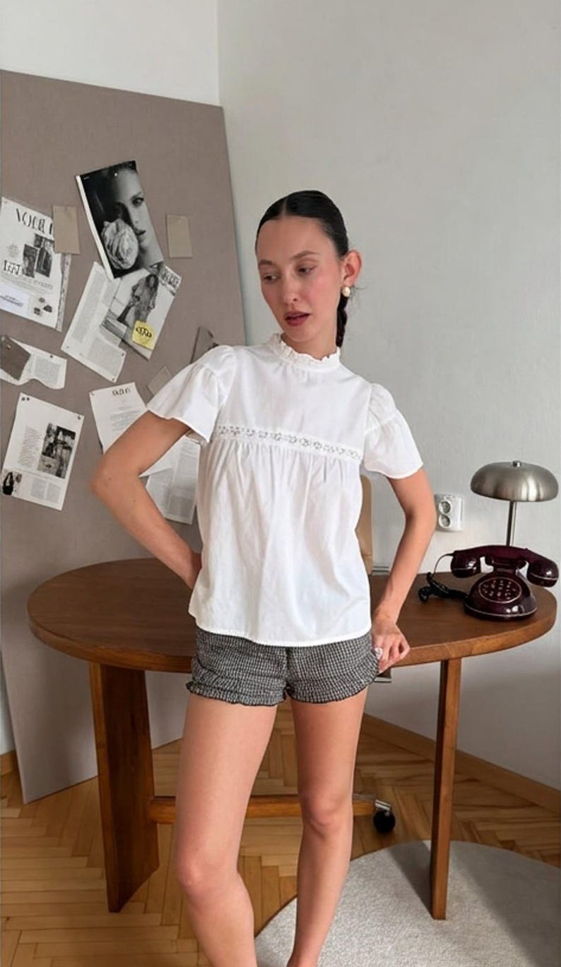 May include: A white short-sleeved blouse featuring a high ruffled collar and a horizontal lace insert across the chest, paired with black and white checkered shorts that have a subtle ruffled hemline along the bottom edge of the garment.