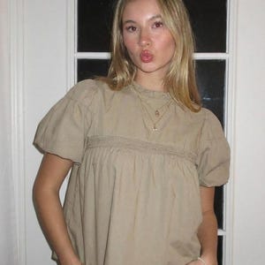 Puede incluir: Una blusa color beige con mangas cortas abullonadas y un panel bordado decorativo en el pecho. La blusa se combina con unos vaqueros azules. Se ven un collar y una pulsera dorados.