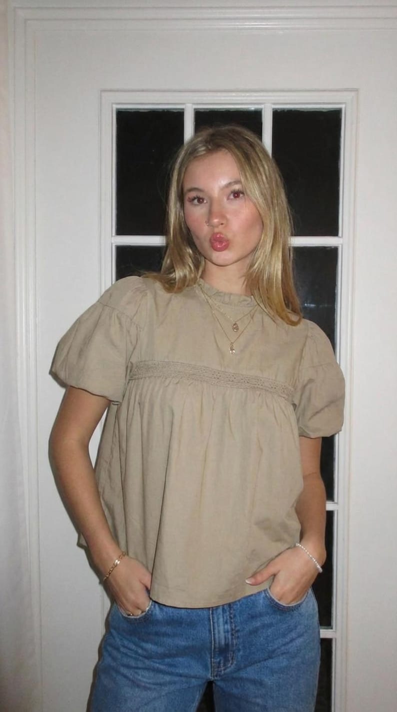 Puede incluir: Una blusa color beige con mangas cortas abullonadas y un panel bordado decorativo en el pecho. La blusa se combina con unos vaqueros azules. Se ven un collar y una pulsera dorados.