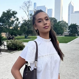 Puede incluir: Una mujer con una blusa blanca con mangas con volantes y un peque&ntilde;o bolso marr&oacute;n. Est&aacute; de pie afuera frente a un parque y un horizonte urbano. Lleva pendientes de aro y tiene el pelo recogido.