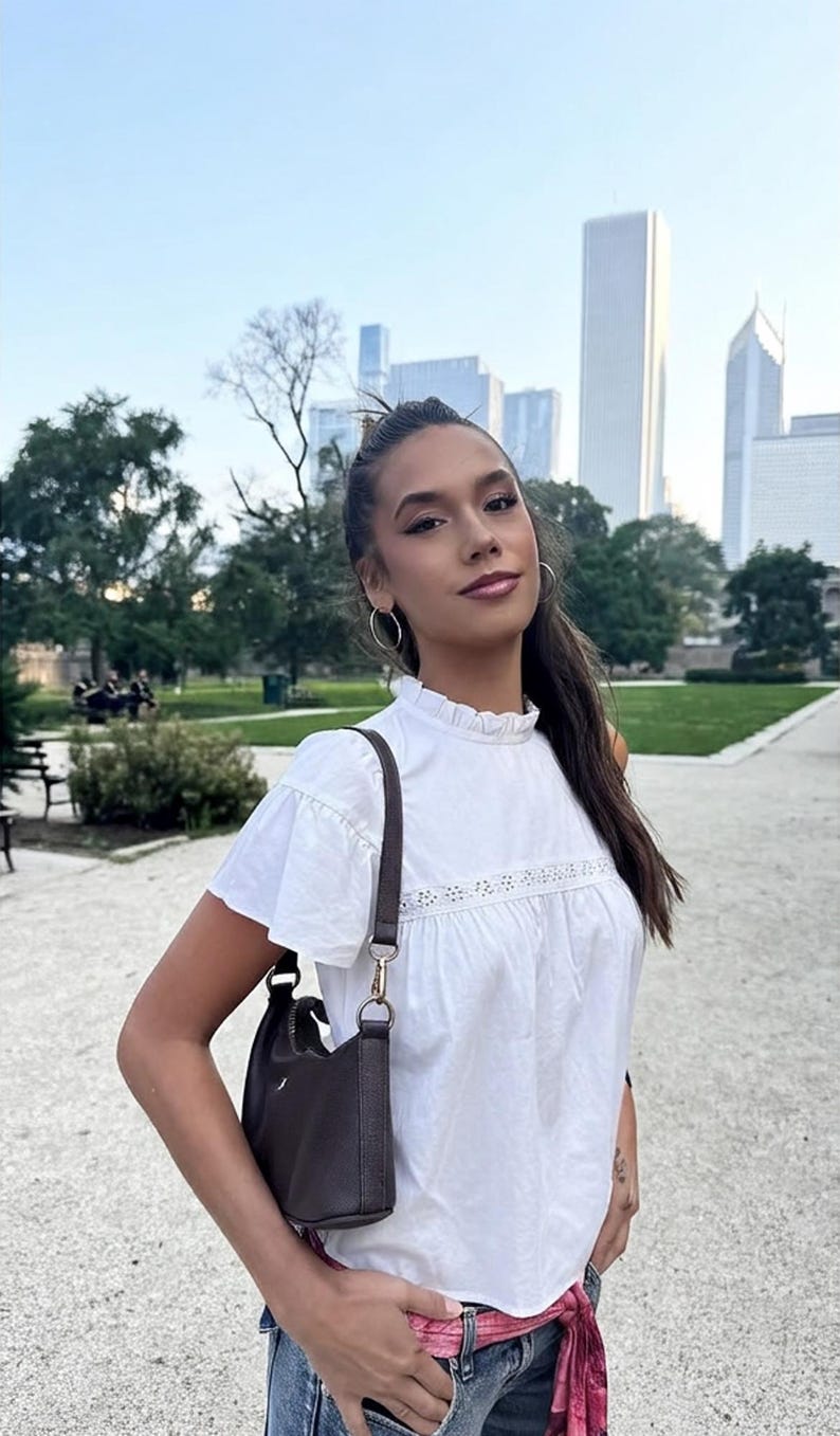 Puede incluir: Una mujer con una blusa blanca con mangas con volantes y un peque&ntilde;o bolso marr&oacute;n. Est&aacute; de pie afuera frente a un parque y un horizonte urbano. Lleva pendientes de aro y tiene el pelo recogido.