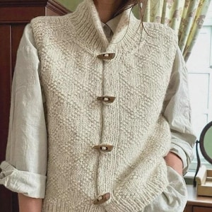 May include: Cream-colored knitted vest with a high collar and wooden toggle closures. The vest features a textured, diamond-patterned design. The garment is worn over a light-colored button-down shirt.