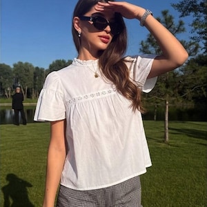 May include: A white short-sleeved blouse featuring a high ruffled collar, a delicate lace trim detail across the chest, and a loose fit, paired with black and white gingham patterned shorts that have a ruffled hemline along the bottom edges.