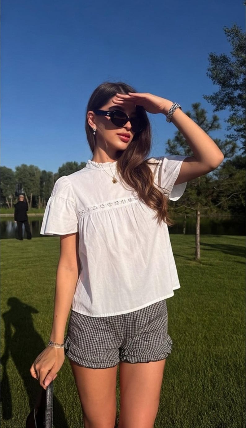 May include: A white short-sleeved blouse featuring a high ruffled collar, a delicate lace trim detail across the chest, and a loose fit, paired with black and white gingham patterned shorts that have a ruffled hemline along the bottom edges.