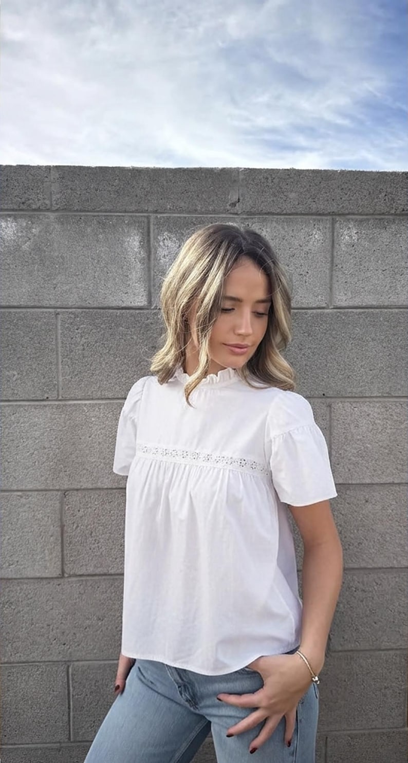 Puede incluir: Una blusa blanca con mangas cortas y cuello con volantes. La blusa tiene un panel bordado decorativo en el pecho. La modelo lleva vaqueros azul claro. El fondo es una pared de ladrillos gris.