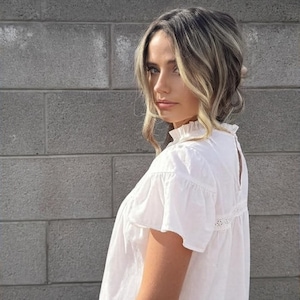 Puede incluir: Una blusa blanca de manga corta con cuello con volantes y detalles de encaje. La blusa se combina con vaqueros azules. El fondo presenta una pared de ladrillos grises y un cielo parcialmente nublado.