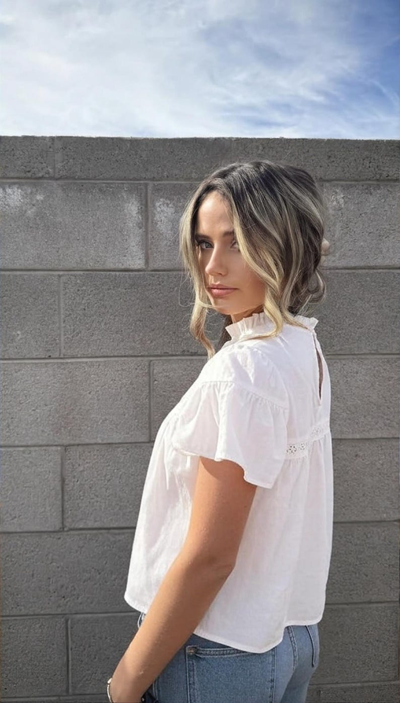 Puede incluir: Una blusa blanca de manga corta con cuello con volantes y detalles de encaje. La blusa se combina con vaqueros azules. El fondo presenta una pared de ladrillos grises y un cielo parcialmente nublado.