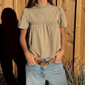 Puede incluir: Una blusa color beige con mangas cortas abullonadas y cuello con volantes. La blusa tiene un detalle de encaje decorativo en el pecho. Se combina con vaqueros azul claro y un cintur&oacute;n de cuero marr&oacute;n. El conjunto es informal y elegante.