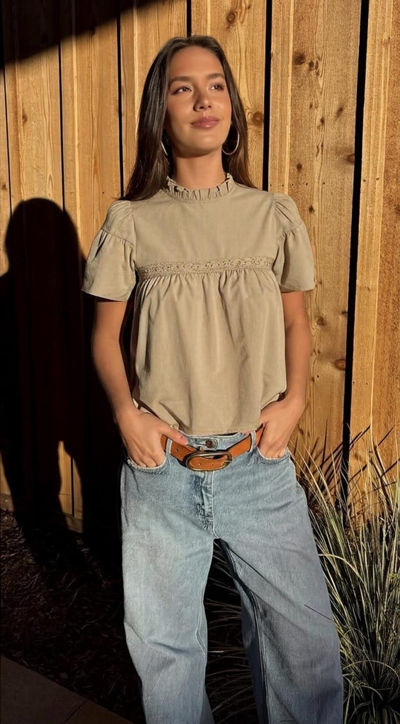 Puede incluir: Una blusa color beige con mangas cortas abullonadas y cuello con volantes. La blusa tiene un detalle de encaje decorativo en el pecho. Se combina con vaqueros azul claro y un cintur&oacute;n de cuero marr&oacute;n. El conjunto es informal y elegante.