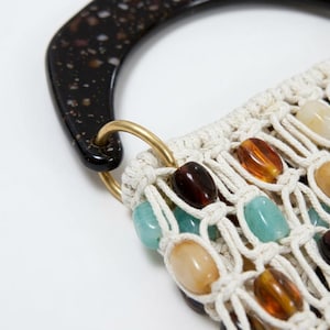 Peut inclure: Un sac à main en macramé composé d'un corps en corde nouée blanc cassé, orné de perles polies dans des tons ambre, brun foncé et turquoise clair. Le sac possède une anse en résine tachetée brun foncé fixée par un anneau en métal doré.