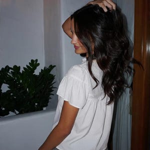 Puede incluir: Una blusa blanca de manga corta con cuello con volantes y un panel de encaje decorativo. La persona lleva un pantal&oacute;n corto a cuadros blanco y negro. La persona se toca el pelo.
