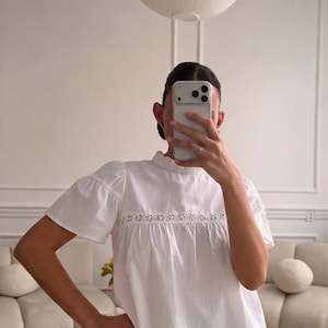 May include: A white short-sleeved blouse featuring a high ruffled collar and a horizontal band of intricate floral eyelet lace across the chest. The garment is paired with dark grey and white checkered shorts that have a ruffled hemline detail.