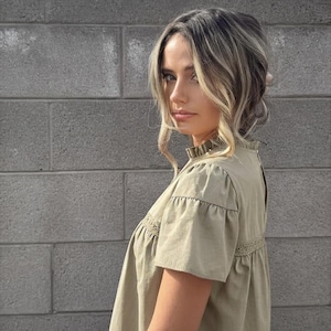 Puede incluir: Top verde oliva de manga corta con cuello con volantes y detalles de encaje. El top tiene un corte holgado y est&aacute; hecho de una tela ligera. La modelo lleva el top con vaqueros azules. El fondo es una pared de ladrillos gris.