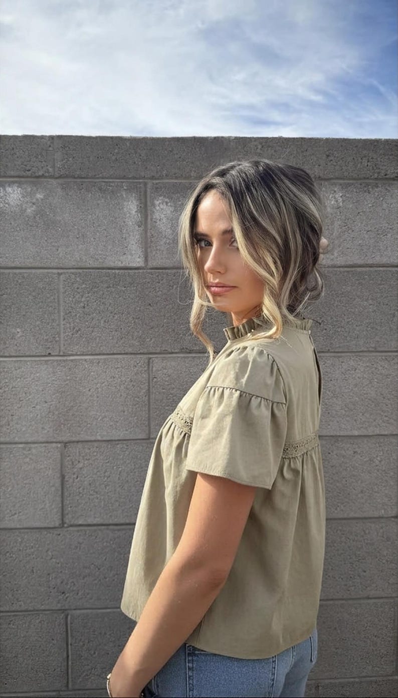 Puede incluir: Top verde oliva de manga corta con cuello con volantes y detalles de encaje. El top tiene un corte holgado y est&aacute; hecho de una tela ligera. La modelo lleva el top con vaqueros azules. El fondo es una pared de ladrillos gris.