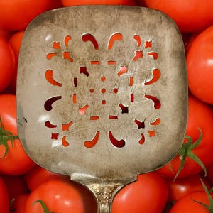 May include: A vintage silver slotted spatula with decorative cutouts, set against a background of ripe red tomatoes. The spatula's handle has an ornate design, and the tomatoes have green stems.