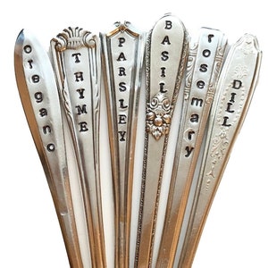 May include: A collection of vintage silver spoons repurposed as garden markers. Each spoon is engraved with a different herb name: oregano, thyme, parsley, basil, rosemary, and dill. The spoons are arranged in a fan shape, showcasing their unique designs.